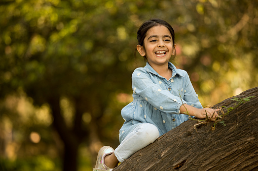 Playful elementary Indian girl laughing and climbing on tree trunk while enjoying weekend in park