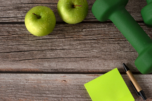 Green dumbbells, apples and blank note on wooden background with copy space. Top view. Weight lifting, fitness and workout, eating healthy diet, training and exercising concept.