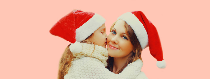 Christmas portrait of little girl child kissing her happy mother in santa red hat on pink background