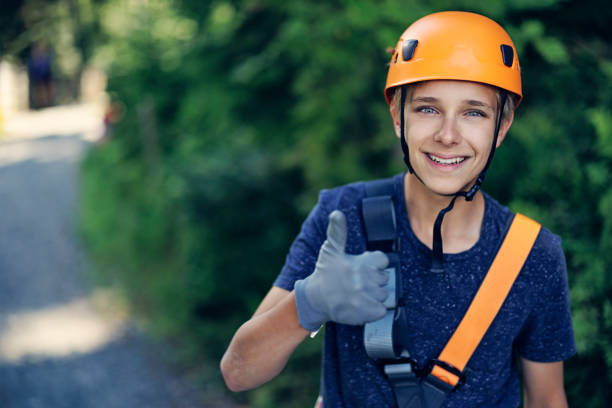 Teenage boy aged 13, wearing helmet and harness is enjoying the outdoors ropes course adventure park. The boy is smiling at the camera and showing thumb up.Canon R5