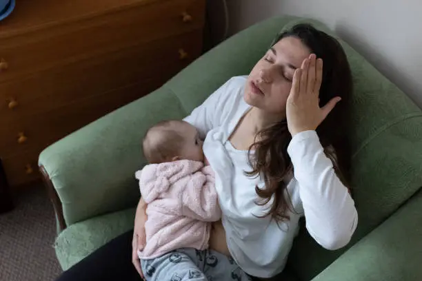 Mother experiences headache while breastfeeding her daughter. Mother sit in the armchair and holds her daughter in her arms while breastfeeding. Baby one year old. The mother takes care of her daughter on maternity leave.