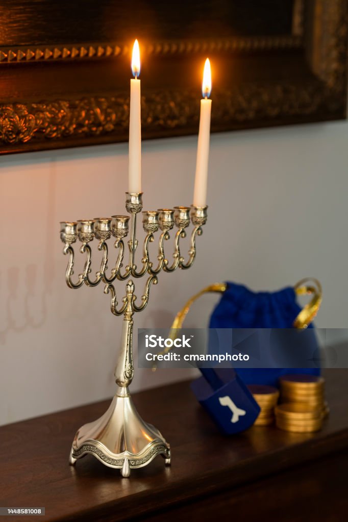 A close-up of a Menorah with one candle and the Shamash lit on a fireplace mantel next to a Dreidel and gold coins for the first night of Hanukkah A close-up of a Menorah burning one candle and Shamash to celebrate the first night of Hanukkah sitting on a stained wooden fireplace mantel with a blue Dreidel and gold coins framed mirror hanging on the wall Atmospheric Mood Stock Photo