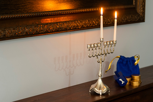 A close-up of a Menorah burning one candle and Shamash to celebrate the first night of Hanukkah sitting on a stained wooden fireplace mantel with a blue Dreidel and gold coins framed mirror hanging on the wall