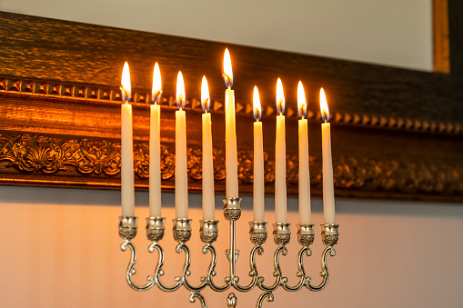 A close-up of a Menorah burning all eight candles and Shamash to celebrate Hanukkah sitting on a stained wooden fireplace mantel with a framed mirror hanging on the wall