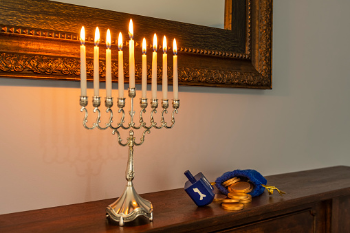 A Menorah burning all eight candles and Shamash to celebrate Hanukkah sitting on a stained wooden fireplace mantel along with a Dreidel and blue pouch with gold coins spilling out.