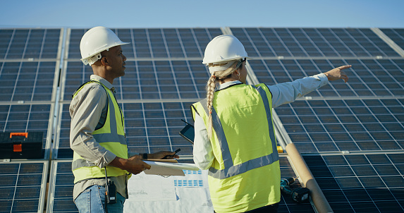 Construction, teamwork and project management for solar panel installation, man and woman meeting with blueprint. Renewable energy, sustainability and solar energy engineering team management on roof