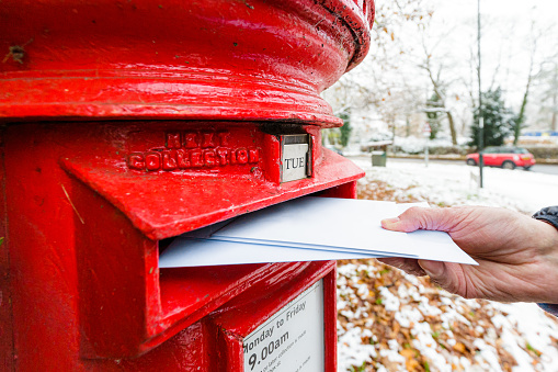 Color image depicting a senior man in his 70s mailing a letter in a traditional red British mailbox while outdoors in winter. There is a layer of snow on top of the mailbox.