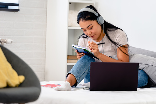 Asian student, a young woman learning the English language over the internet at home. Using headphones and a laptop computer, attending an online class