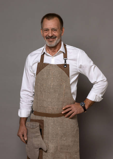 A mature man in a chef's apron with an oven mitt is standing and smiling. A mature man in a chef's apron with an oven mitt is standing and smiling. Isolated background. snakes beard stock pictures, royalty-free photos & images