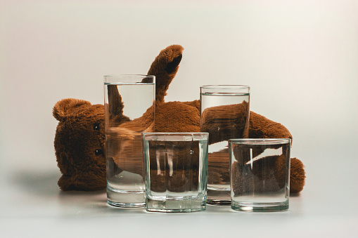 Still life with a brown teddy bear on a white background, in front of which are glass glasses with water, creating an illusion.