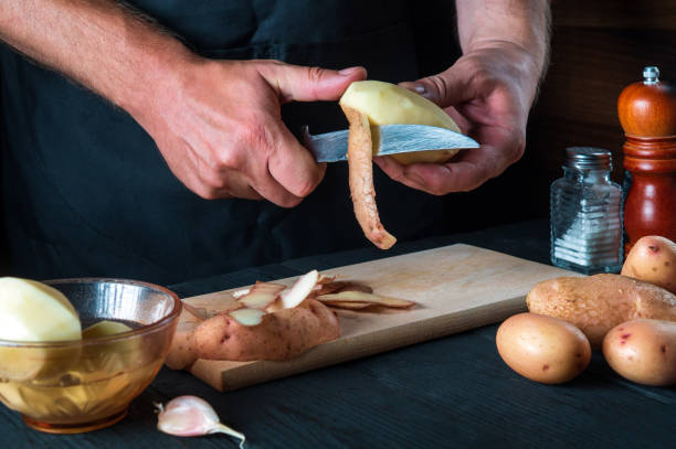 istock Chef cleans potatoes for mashed potatoes, stews, roasted and with vegetables. For dishes with potatoes. On a black background, the concept of the menu, cooking, healthy eating 1448596961