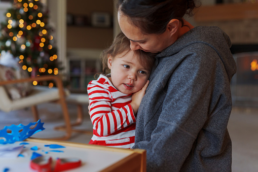 An adorable Eurasian toddler girl takes a break from helping her mom with a Christmas themed art and craft project to give her mom a big hug. The family is at home in the living room and a Christmas tree strung with lights twinkles in the background