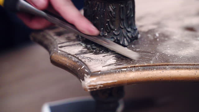 Carpentry expert renovating vintage wooden table with bronze legs in workshop, close up shot of worker hand using industrial chisel tool to remove old colour paint from wooden surface, destructed wood surface in hands of skill carpenter, furnitures renov