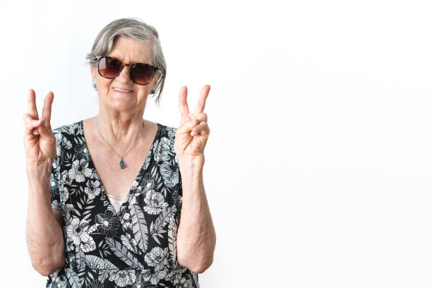 istock Smiling elderly woman wearing sunglasses and doing peace symbol while looking at camera over white background 1448602853
