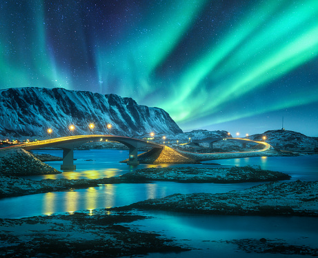 Bridge and northern lights over snowy mountains. Lofoten islands, Norway. Aurora borealis and reflection in water. Winter landscape with starry sky, polar lights, rocks, road, sea, city illumination