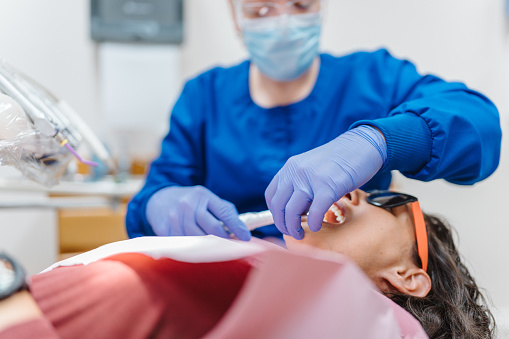 A multiracial female patient lies in a dentist's chair and opens her mouth so her dentist can examine and clean her teeth during a routine appointment. Selective focus on the dentist's hands holding medical equipment.