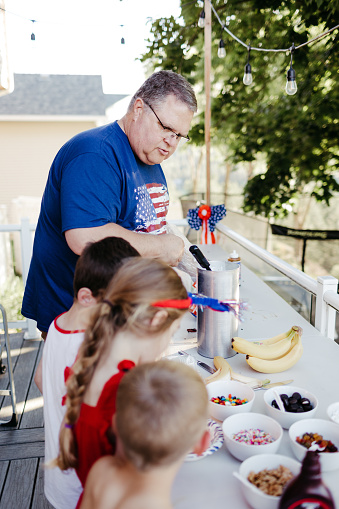 A mature adult man wearing an American Flag t-shirt helps his elementary age grandkids make ice cream sundaes while celebrating the Fourth of July outside on the back patio on a hot summer afternoon.