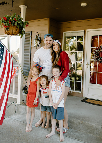 Portrait of a happy husband and wife and their three young children smiling directly at the camera while posing on the front porch of their home on USA Independence Day.