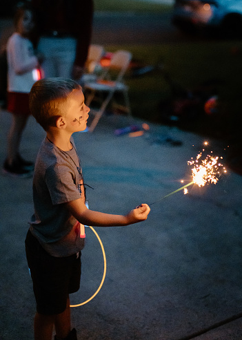 A young boy stands in his family's driveway at night and holds a lit sparkler firework while celebrating the Fourth of July on a hot summer evening.
