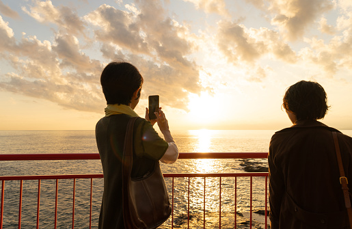 Japanese mother and daughter sightseeing in Japan.\nBack view of mother and daughter taking a picture of the sunset with a smart phone.\nActual mother and daughter.\nEnoshima Island, Kanagawa, Japan.