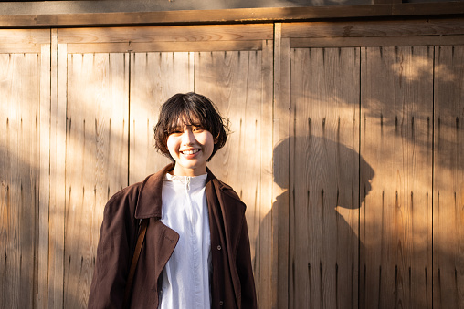 Portrait of an Asian woman.\nTaken in front of a wooden wall.\nShe is looking at the camera and smiling happily.\nJapanese woman in her 20s.