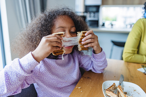 A portrait of a cute little girl playing with her food while having breakfast with her parents. She is breaking the cheesy toast sandwich in half and letting the melted cheese drag as she is pulling the bread apart. The adorable daughter is wearing a purple pajama and sitting by a wooden dining table in a modern apartment.