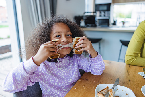 A portrait of a cute young daughter breaking a cheesy toast sandwich in half. She is smiling and looks happy while she is playing with her food. She is sitting by a wooden table and has a white plate in front of her. She is located in a beautiful kitchen with modern interior and big windows. The young girl is wearing a cute purple pajama.