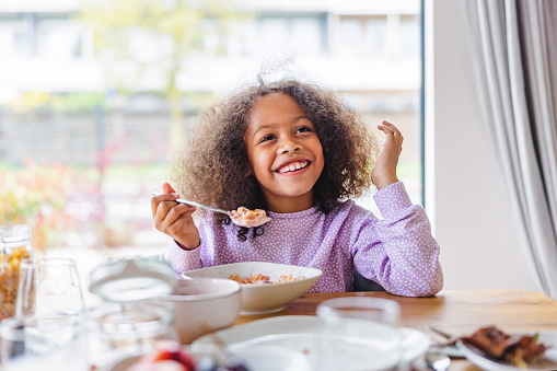 Happy adorable black girl sitting by a wooden table full of food and eating a bowl of cereal for breakfast in the morning. She is smiling. The girl has long curly hair and is wearing purple pajamas. She is located in a modern apartment with big windows and a nice view.
