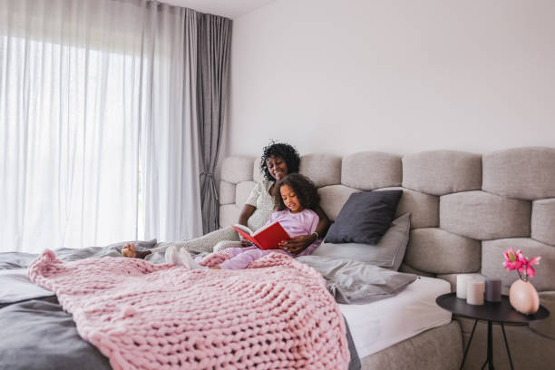 istock Black Little Daughter Learning How To Read While Lying In Bed Next To Her Supportive Black Pregnant Mother 1448623422