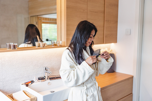 Attractive caucasian female with long dark hair brushing her hair with a hair brush after taking a hot shower. She is wearing a white bathrobe and is standing in a beautiful modern bathroom. There is a big mirror and a white sink with wooden cabinets behind her. The beautiful led lights create a cosy environment.
