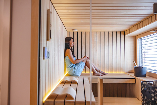 Beautiful young adult caucasian female lying on the bench with her knees bent and leaning against the wooden wall in a small cosy sauna in a spa hotel. She is alone and looking through a big widow with blinds. The is wearing just a towel and looking forward. There are beautiful cosy lights in the sauna.