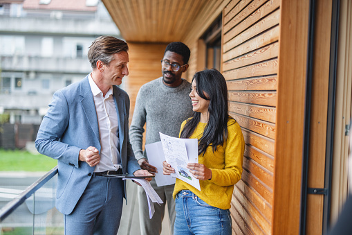 An adult caucasian male real estate agent talking to his clients while giving them a tour of the house. They are standing on the balcony while he is showing the the view. The agent is dressed in a fancy suit while the diverse couple is dressed in casual sweaters and jeans. The couple is interested in buying the house. They are holding some papers in their hands.