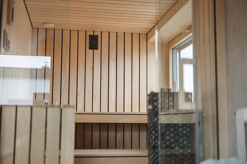 A beautiful cosy wooden sauna located in a modern apartment. It has wooden panel and a place to sit and lie. It looks relaxing and has a lot of natural light due to the big window in the sauna. The sauna's doors are glass making it look luxurious.
