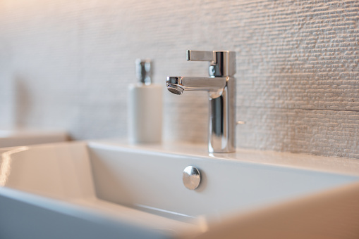 A close up of a white cermic bathroom sink with aluminium tap and silver details. There is a white and silver container for liquid soap on the sink. The sink looks very clean and shiny.