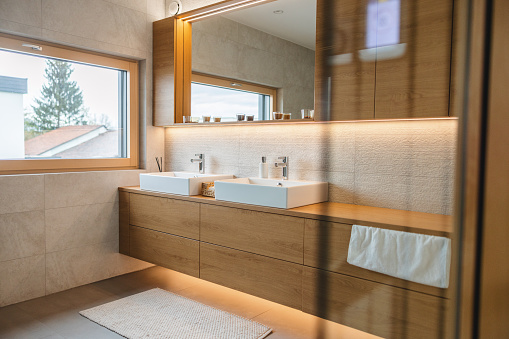 A modern bathroom with two sinks and a big mirror. The interior of the bathroom is modern and cosy. It's brown and beige. There are wooden details and bathroom cabinets and stone tiles. The bathroom is illuminated with beautiful led lights hidden behind the mirror and under the cabinets. There are some small candles to make the bathroom feel more homey.