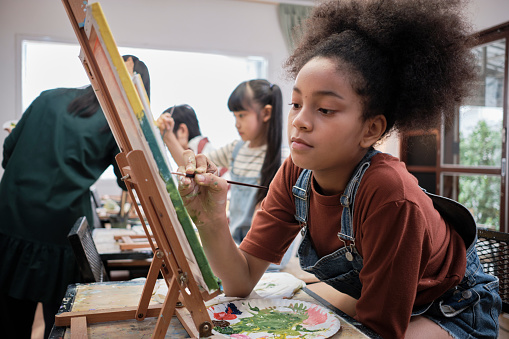 African American girl concentrates on acrylic color picture painting on canvas with students group in art classroom, creative learning with talents and skills in elementary school studio education.