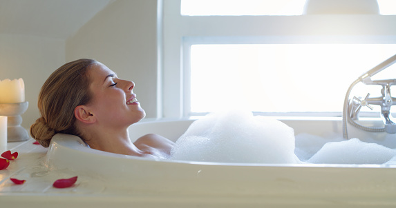 Rose, relax and woman in a bubble bath with bubbles and a candle on her luxury vacation holiday in a resort's bathroom. Hospitality, alone and happy girl cleaning in soap water with foam at a hotel