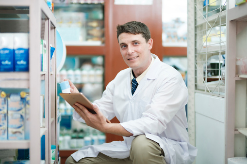 Male smart young-adult pharmacist in professional white gown looking and checking stock inventory in modern pharmacy, drugstore indoor using tablet. Pharmacy, medicine, and healthcare concept.
