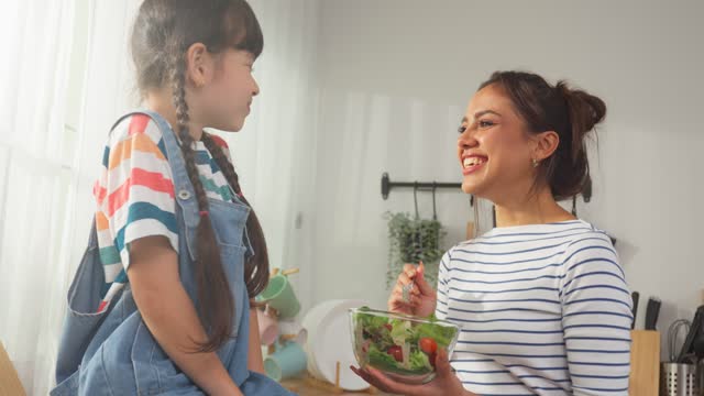 Asian mother teach and motivate young girl child eat green vegetable. Adorable little kid child feel happy and relax, enjoy eat salad and healthy foods for health care and wellness in kitchen at home.