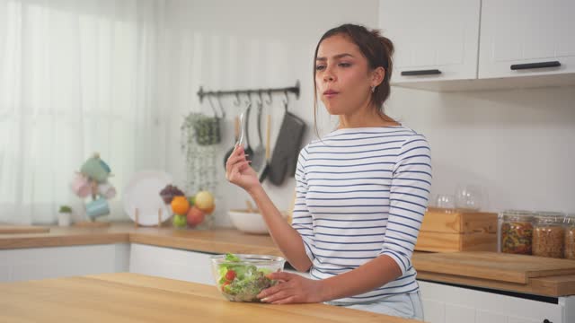 Young Caucasian girl eating green healthy salad in kitchen at home. Attractive beautiful vegetarian woman feeling happy and enjoy healthy foods for breakfast to lose weight and health care in house.