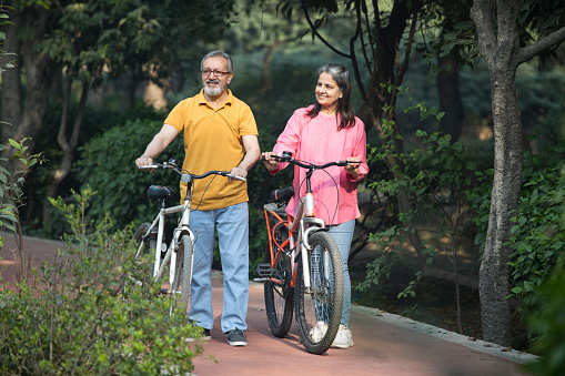 Carefree senior couple walking with bicycles on road amidst plants in park during weekend