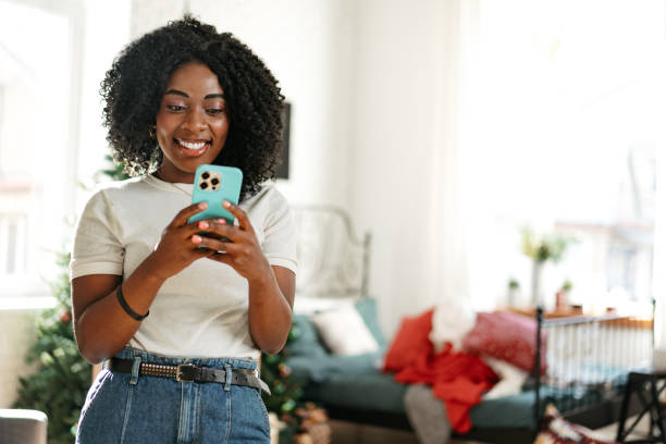 Smiling african woman sitting on couch at home and using smartphone