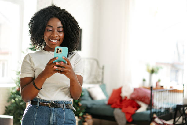 Smiling african woman sitting on couch at home and using smartphone stock photo