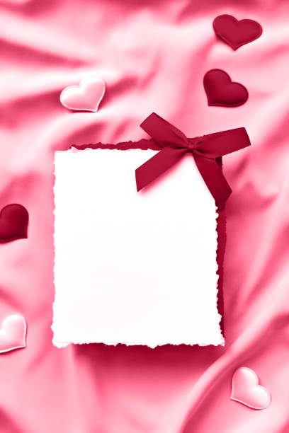 An empty white sheet of paper with a red bow and decorative hearts on pink silk. Flat lay, place for text. vertical image. Demonstrating the colors of 2023 Viva Magenta.