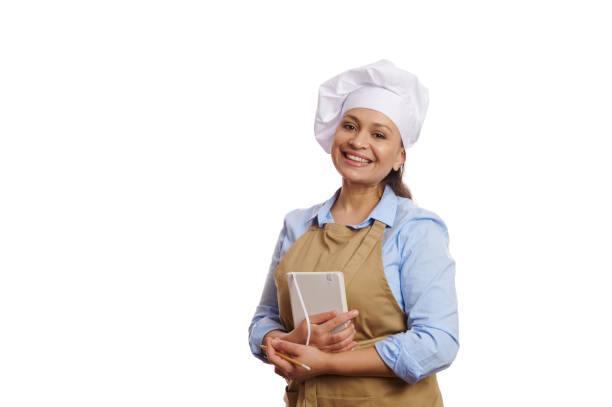 istock Charming female chef confectioner in apron, holding her notepad and cutely smiling looking at camera, white background 1448680762