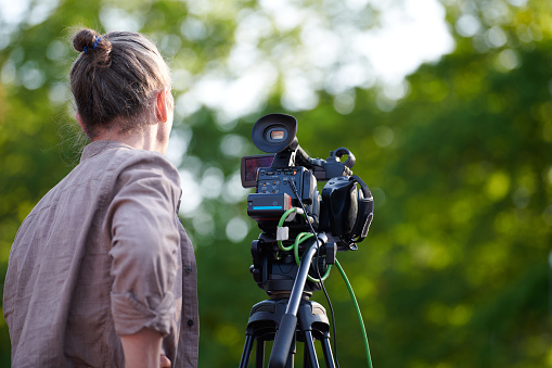 Rear view to man videographer recording outdoor movie about nature, back view to TV channel male crew with TV camera, green background. Long haired man with professional camera records video for TV