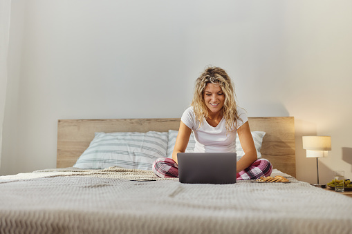 Young happy woman reading an e-mail on a computer while relaxing during morning on a bed. Copy space.