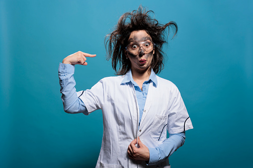 Insane and funny looking chemist gesturing craziness by twisting finger at temple while having dirty face and messy hair. Amusing scientist with wacky appearance looking at camera on blue background.