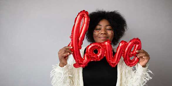 A banner with an African-American happy girl on a gray background, holding a balloon with the inscription Love. A woman's smile when she is in love. A concept for All Lovers