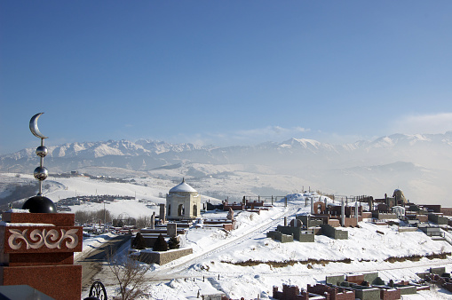 Cemetery with some graves a bit outside of Almaty City. The image shows the cemetery with snow covered mountain peaks in the d during winter season.